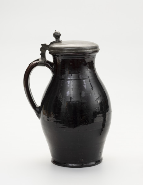 Dzban z pokrywą - ujęcie z tyłu; Wysmukły dzban o lekko wyodrębnionej podstawie. Największa szerokość brzuśca przypada w połowie wysokości naczynia. Brzusiec łagodnym przewężeniem przechodzi w szeroką szyjkę; miejsce przejścia zaznaczone dookolną linią. Szyjka łagodnie przechodzi w wywinięty na zewnątrz wylew o zaoblonym rancie. Z lewej szerokie ucho, taśmowe, zaczepione pomiędzy dolną częścią szyjki, a  brzuścem poniżej najszerszej jego części. Na szczycie ucha obejma ze skierowaną pionowo do góry, ozdobnie profilowana kolumienką zaopatrzoną w zawias, służący do przymocowania pokrywy, szczelnie zamykającej wylew. Pokrywka z bolcem w postaci kolumienki zakończonej kopułką z małą kulką na szczycie, tworzące paluch do jej podnoszenia. Na pokrywie proste liniowo-koncentryczne zdobienie uzyskane prawdopodobnie cyzelowaniem. Dzban  w całości pokryty szkliwem, w środku cieńszą warstwą a na zewnątrz grubszą. Widoczne ślady po odrutowaniu, utrzymującym w całości pękniętą konstrukcję.