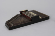 Cytra klawiszowa (autoharp) - ujęcie ze skosu; Cytra klawiszowa (autoharp) z płaskim pudłem rezonansowym wykonanym z drewna polichromowanego o kształcie przypominającym trapez z jednym ściętym rogiem. Wzdłuż najkrótszego boku oraz boku odchodzącego od niego pod kontem rozwartym, szereg metalowych kołków, do których przymocowanych jest 25, diatonicznie strojonych strun, ułożonych od najgrubszej – dolnej – do najcieńszej górnej strony instrumentu; ostatnie dwie są zerwane. Z prawej strony kołków znajdują się dwie, zestawione pod kątem rozwartym, wąskie, metalowe blaszki z niewielkimi bolcami, stanowiące wsparcie i gwarantujące właściwy rozstaw strun. Podobna blaszka zamontowana jest wzdłuż dolnej krawędzi instrumentu. Struny przechodzą przez znajdujące się w niej rowki. Bezpośrednio nad strunami, prostopadle względem nich, znajduje się sześć metalowych prętów zamontowanych pomiędzy dwoma drewnianymi mostkami. Osadzone na sprężynach pręty wprawiane są w ruch przez grającego za pomocą znajdujących się w ich górnej części przycisków. Powoduje to dociśnięcie do struny mocowanych na prętach klawiszy z filcowymi podkładkami. Na górnym mostku, na przykręconej za pomocą dwóch śrub plakietce z papieru welinowego, znajdują się oznaczenia akordów. Druga plakietka, z zapisem nut w formie literowej i na pięciolinii oraz ciągiem cyfr rzymskich i arabskich, umieszczona jest bezpośrednio na pudle rezonansowym, z prawej strony mechanizmu klawiszowego. Cytra ozdobiona jest w lewej części kalkomanią przedstawiającą ptaszka, prawdopodobnie wróbla, siedzącego na ukwieconej gałązce.