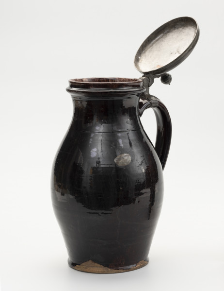 Dzban z pokrywą - ujęcie ze skosu; Wysmukły dzban o lekko wyodrębnionej podstawie. Największa szerokość brzuśca przypada w połowie wysokości naczynia. Brzusiec łagodnym przewężeniem przechodzi w szeroką szyjkę; miejsce przejścia zaznaczone dookolną linią. Szyjka łagodnie przechodzi w wywinięty na zewnątrz wylew o zaoblonym rancie. Ucho szerokie, taśmowe, zaczepione pomiędzy dolną częścią szyjki, a  brzuścem poniżej najszerszej jego części. Na szczycie ucha obejma ze skierowaną pionowo do góry, ozdobnie profilowana kolumienką zaopatrzoną w zawias, służący do przymocowania pokrywy, szczelnie zamykającej wylew. Pokrywka otwarta z bolcem w postaci kolumienki zakończonej kopułką z małą kulką na szczycie, tworzące paluch do jej podnoszenia. Dzban  w całości pokryty szkliwem. Naczynie z wyraźnymi śladami naprawy: dziura w brzuścu zalepiona cynową, lutowana łatą, widoczne ślady po odrutowaniu, utrzymującym w całości pękniętą konstrukcję.