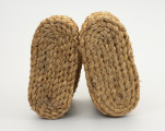 Buty słomiane - ujęcie z dołu; Buty słomiane wykonane techniką plecionkarską. Buty o gładkiej podeszwie (bez obcasa). Poszczególne elementy spirali warkocza  zszywane sznurkiem konopnym. Buty słomiane stosowane były do ocieplenia nóg w trakcie zimowych prac gospodarczo - rolniczych, jak np. w czasie młocki cepami.