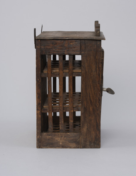 Szafka do przechowywania jajek - ujęcie z lewego boku; Szafka do przechowywania jajek o konstrukcji ramowej w kształcie prostopadłościanu. Płycina ścianek bocznych  wypełniona trzema pionowymi szczebelkami. Drzwiczki zamykane na zapadkę uruchamianą klamką. Wewnątrz trzy półki z wyciętymi otworami do wkładania jaj. Na desce zamykającej szafkę od góry przybita ozdobnie fazowana listwa. Na górze dwa haczyki do zawieszania.