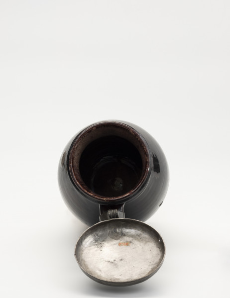 Dzban z pokrywą - ujęcie z góry; Ujęcie wnętrza dzbana. Brzusiec łagodnym przewężeniem przechodzi w szeroką szyjkę. Przy szyjce zawias, służący do przymocowania pokrywy, szczelnie zamykającej wylew. Otwarta pokrywka z bolcem. Dzban pokryty szkliwem, w środku cieńszą warstwą a na zewnątrz grubszą.