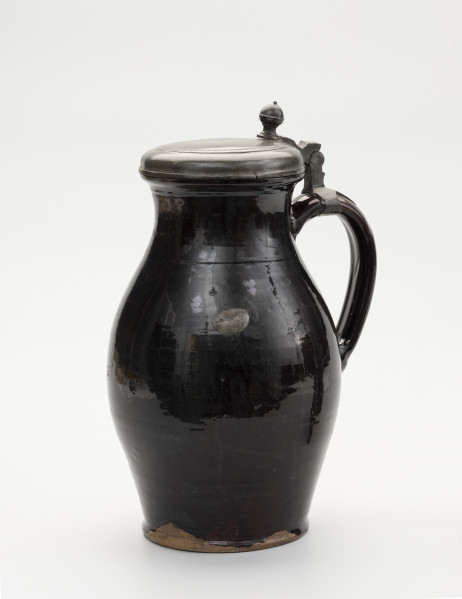 Dzban z pokrywą - ujęcie ze skosu; Wysmukły dzban o lekko wyodrębnionej podstawie. Największa szerokość brzuśca przypada w połowie wysokości naczynia. Brzusiec łagodnym przewężeniem przechodzi w szeroką szyjkę; miejsce przejścia zaznaczone dookolną linią. Szyjka łagodnie przechodzi w wywinięty na zewnątrz wylew o zaoblonym rancie. Ucho szerokie, taśmowe, zaczepione pomiędzy dolną częścią szyjki, a  brzuścem poniżej najszerszej jego części. Na szczycie ucha obejma ze skierowaną pionowo do góry, ozdobnie profilowana kolumienką zaopatrzoną w zawias, służący do przymocowania pokrywy, szczelnie zamykającej wylew. Pokrywka z bolcem w postaci kolumienki zakończonej kopułką z małą kulką na szczycie, tworzące paluch do jej podnoszenia. Na pokrywie proste liniowo-koncentryczne zdobienie uzyskane prawdopodobnie cyzelowaniem. Dzban  w całości pokryty szkliwem, w środku cieńszą warstwą a na zewnątrz grubszą. Naczynie z wyraźnymi śladami naprawy: dziura w brzuścu zalepiona cynową, lutowana łatą, widoczne ślady po odrutowaniu, utrzymującym w całości pękniętą konstrukcję. Od dołu widoczne ubytki szkliwa.