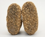 Buty słomniane - ujęcie z dołu; Buty wykonane ze słomy (prawdopodobnie żytniej) techniką spiralną, wyrób plecionkarski, jednostkowy. Podeszwa płaska. Wykonane z warkocza plecionego z powróseł słomianych różnej grubości, początek zawiązuje się  słomą lub sznurkiem i pelcie się rękoma warkocz, dokładając co pewien czas nowe pasmo słomy. Poszczególne rzędy, z których wypleciono buty powiązane są ze sobą za pomocą sznurka. Buty takie służyły jako ocieplacz nóg i izolator przed zimnym podłożem w trakcie zimowych prac gospodarskich.