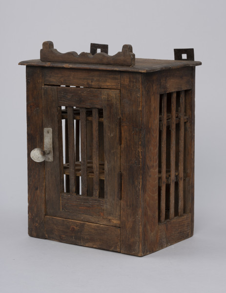 Szafka do przechowywania jajek - ujęcie ze skosu; Szafka do przechowywania jajek o konstrukcji ramowej w kształcie prostopadłościanu. Płycina jednoskrzydłowych drzwi oraz ścianki boczne  otwarte, wypełnione trzema pionowymi szczebelkami. Drzwiczki zamykane na zapadkę uruchamianą klamką. Wewnątrz półki z wyciętymi otworami do wkładania jaj. Na desce zamykającej szafkę od góry przybita ozdobnie fazowana listwa. Na górze przybite dwa haczyki do zawieszania.