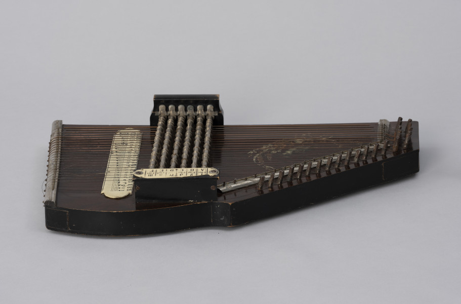 Cytra klawiszowa (autoharp) - ujęcie z boku; Cytra klawiszowa (autoharp) z płaskim pudłem rezonansowym wykonanym z drewna polichromowanego o kształcie przypominającym trapez z jednym ściętym rogiem.  Wzdłuż najkrótszego boku oraz boku odchodzącego od niego pod kontem rozwartym, szereg metalowych kołków, do których przymocowanych jest 25, diatonicznie strojonych strun, ułożonych od najgrubszej – od góry – do najcieńszej u dołu; ostatnie dwie na dole są zerwane. Na lewo od kołków znajdują się dwie, zestawione pod kątem rozwartym, wąskie, metalowe blaszki z niewielkimi bolcami, stanowiące wsparcie i gwarantujące właściwy rozstaw strun. Podobna blaszka zamontowana jest wzdłuż dolnej krawędzi instrumentu. Struny przechodzą przez znajdujące się w niej rowki, zaginają pod kątem 90 stopni i mocowane są w znajdujących się w bocznej ściance instrumentu bolcach. Bezpośrednio nad strunami, prostopadle względem nich, znajduje się sześć metalowych prętów zamontowanych pomiędzy dwoma drewnianymi mostkami. Na jednym z mostków, na przykręconej za pomocą dwóch śrub plakietce z papieru welinowego, znajdują się oznaczenia akordów. Druga plakietka, z zapisem nut w formie literowej i na pięciolinii oraz ciągiem cyfr rzymskich i arabskich, umieszczona jest bezpośrednio na pudle rezonansowym. Cytra ozdobiona jest z prawej strony kalkomanią.