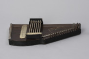 Cytra klawiszowa (autoharp) - ujęcie z boku; Cytra klawiszowa (autoharp) z płaskim pudłem rezonansowym wykonanym z drewna polichromowanego o kształcie przypominającym trapez z jednym ściętym rogiem.  Wzdłuż najkrótszego boku oraz boku odchodzącego od niego pod kontem rozwartym, szereg metalowych kołków, do których przymocowanych jest 25, diatonicznie strojonych strun, ułożonych od najgrubszej – od góry – do najcieńszej u dołu; ostatnie dwie na dole są zerwane. Na lewo od kołków znajdują się dwie, zestawione pod kątem rozwartym, wąskie, metalowe blaszki z niewielkimi bolcami, stanowiące wsparcie i gwarantujące właściwy rozstaw strun. Podobna blaszka zamontowana jest wzdłuż dolnej krawędzi instrumentu. Struny przechodzą przez znajdujące się w niej rowki, zaginają pod kątem 90 stopni i mocowane są w znajdujących się w bocznej ściance instrumentu bolcach. Bezpośrednio nad strunami, prostopadle względem nich, znajduje się sześć metalowych prętów zamontowanych pomiędzy dwoma drewnianymi mostkami. Na jednym z mostków, na przykręconej za pomocą dwóch śrub plakietce z papieru welinowego, znajdują się oznaczenia akordów. Druga plakietka, z zapisem nut w formie literowej i na pięciolinii oraz ciągiem cyfr rzymskich i arabskich, umieszczona jest bezpośrednio na pudle rezonansowym. Cytra ozdobiona jest z prawej strony kalkomanią.