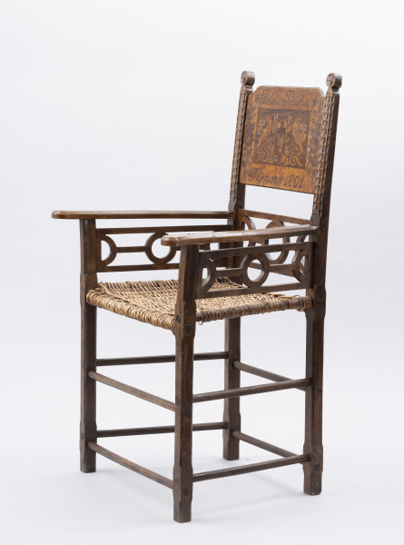 Fotel - ujęcie z boku; Fotel - krzesło z podłokietnikami, wyrób rzemieślniczy stolarski, wykonawca nieznany. Mebel o konstrukcji ramowej z pionowo wznoszącym się oparciem. Nogi proste o przekroju prostokątnym, łączone dwiema toczonymi podpórkami, przednie z delikatnym wzorem geometrycznym wykonanym techniką snycerską. Siedzisko proste, wyplatane wikliną. Pomiędzy siedziskiem, dolną częścią oparcia i podłokietnikami ażurowo ozdobnie wycięta listwa ( motyw koła) będąca zarazem elementem konstrukcji. Podłokietniki na końcach lekko wywinięte na zewnątrz. Rama oparcia na obu listwach ze snycerską dekoracją w postaci stylizowanych kwiatów ( ?) i górnym zwieńczeniem w kształcie okręgu z rytym krzyżem. W ramę, w jej górnej części wpuszczona deska, jej górne krawędzie lekko wycięte na zewnętrznych rogach. Powierzchnia deski, z wyraźnie widoczną strukturą drewna, bogato zdobiona intarsją w dwóch odcieniach drewna. W środkowej części zaznaczony ciemniejszym kolorem drewna prostokąt, w środku którego symetrycznie umieszczone ptaki ( pawie ?)siedzące na stylizowanych gałęziach tulipanów i słoneczników (?). Nad prostokątem napis: Peter Buhr, pod nim: Anno 1801, po bokach motywy florystyczne.