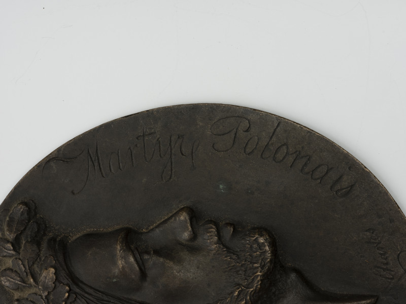 Medalion z wizerunkiem Karola Levittoux - detal; Okrągły medalion jednostronny. Widoczny fragment głowy Karola Levittoux w wieńcu dębowym w prawo. W otoku wygrawerowany napis: Martyre Polonais [polski męczennik].