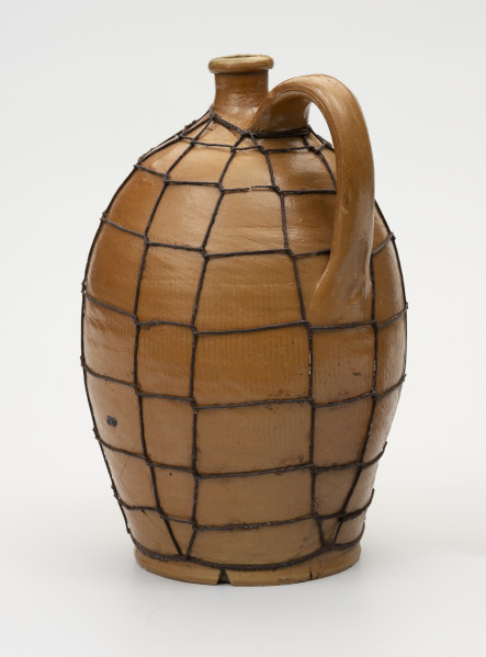 Butla z odrutowaniem - ujęcie ze skosu; Kamionkowa butla o owalnym kształcie i wyraźnie wyodrębnionym dnie. Brzusiec w dolnej części wyraźnie węższy, o największym rozchyleniu powyżej połowy wysokości, w górnej partii przechodzi bezpośrednio w szyjkę. Szyjka krótka, wąska, wyraźnie wyodrębniona z pogrubionym wylewem. Ucho taśmowe, esowato wygięte z górnym punktem zaczepienia nieco poniżej miejsca przejścia brzuśca w szyjkę i dolnym - powyżej połowy wysokości naczynia. Glazura na zewnątrz brązowa. Butla odrutowana cienkim drutem dla wzmocnienia uszkodzonego naczynia. Pęknięcie biegnie z lewej strony od stopy do 1/3 wysokości naczynia.