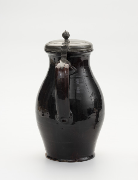 Dzban z pokrywą - ujęcie z drugiego boku; Wysmukły dzban o lekko wyodrębnionej podstawie. Największa szerokość brzuśca przypada w połowie wysokości naczynia. Brzusiec łagodnym przewężeniem przechodzi w szeroką szyjkę; miejsce przejścia zaznaczone dookolną linią. Szyjka łagodnie przechodzi w wywinięty na zewnątrz wylew o zaoblonym rancie. Z przodu ucho szerokie, taśmowe, zaczepione pomiędzy dolną częścią szyjki, a  brzuścem poniżej najszerszej jego części. Na szczycie ucha obejma ze skierowaną pionowo do góry, ozdobnie profilowana kolumienką zaopatrzoną w zawias, służący do przymocowania pokrywy, szczelnie zamykającej wylew. Pokrywka z bolcem w postaci kolumienki zakończonej kopułką z małą kulką na szczycie, tworzące paluch do jej podnoszenia. Na pokrywie proste liniowo-koncentryczne zdobienie uzyskane prawdopodobnie cyzelowaniem. Dzban  w całości pokryty szkliwem, w środku cieńszą warstwą a na zewnątrz grubszą. Widoczne ślady po odrutowaniu, utrzymującym w całości pękniętą konstrukcję.