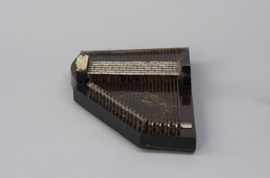 Cytra klawiszowa (autoharp) - ujęcie z drugiego boku; Cytra klawiszowa (autoharp) z płaskim pudłem rezonansowym wykonanym z drewna polichromowanego o kształcie przypominającym trapez z jednym ściętym rogiem. Wzdłuż najkrótszego boku oraz boku odchodzącego od niego pod kontem rozwartym, szereg metalowych kołków, do których przymocowanych jest 25, diatonicznie strojonych strun, ułożonych od najgrubszej – z prawej – do najcieńszej z lewej strony instrumentu; ostatnie dwie są zerwane. Powyżej kołków znajdują się dwie, zestawione pod kątem rozwartym, wąskie, metalowe blaszki z niewielkimi bolcami, stanowiące wsparcie i gwarantujące właściwy rozstaw strun. Podobna blaszka zamontowana jest wzdłuż górnej krawędzi instrumentu. Struny przechodzą przez znajdujące się w niej rowki. Bezpośrednio nad strunami, prostopadle względem nich, znajduje się sześć metalowych prętów zamontowanych pomiędzy dwoma drewnianymi mostkami. Osadzone na sprężynach pręty wprawiane są w ruch przez grającego za pomocą znajdujących się w ich górnej części przycisków. Powoduje to dociśnięcie do struny mocowanych na prętach klawiszy z filcowymi podkładkami. Na mostku z lewej, na przykręconej za pomocą dwóch śrub plakietce z papieru welinowego, znajdują się oznaczenia akordów. Cytra ozdobiona jest kalkomanią.