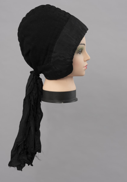 Czepek damski - ujęcie z boku; Czepek damski, codzienny. Wykonany z tkanin produkcji fabrycznej (wełna, atłas) i białego, lnianego płótna wykonanego na krosnach tkackich splotem płóciennym prostym. Główka czepca z tkaniny wełnianej, czarnej, wokół główki naszyta czarna atłasowa tkanina. Z tyłu dekoracyjna kokarda z atłasowej wstążki. Czepek zszyty z dwóch kawałków, szew wzdłuż pionowej linii głowy, ściągany nad karkiem nicią. Główka wyraźnie zaokrąglona, zachodząca wyraźnie na uszy. Od środka czepek podbity lnianym, białym płótnem i wypełniony grubym usztywniającym materiałem. Przez wszystkie warstwy tkanin przechodzą w wielu miejscach przeszycia białymi i czarnymi nićmi tworzącymi ich złączenie  jako przepikowanie tkaniny.  

Literatura: 