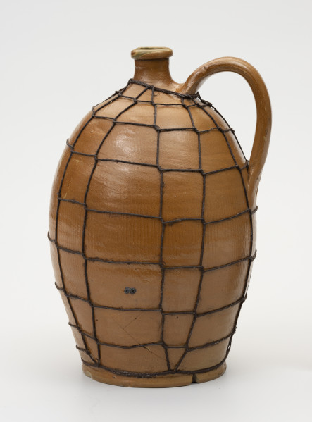 Butla z odrutowaniem - ujęcie z tyłu; Kamionkowa butla o owalnym kształcie i wyraźnie wyodrębnionym dnie. Brzusiec w dolnej części wyraźnie węższy, o największym rozchyleniu powyżej połowy wysokości, w górnej partii przechodzi bezpośrednio w szyjkę. Szyjka krótka, wąska, wyraźnie wyodrębniona z pogrubionym wylewem. Z prawej strony ucho taśmowe, esowato wygięte z górnym punktem zaczepienia nieco poniżej miejsca przejścia brzuśca w szyjkę i dolnym - powyżej połowy wysokości naczynia. Glazura na zewnątrz brązowa. Butla odrutowana cienkim drutem dla wzmocnienia uszkodzonego naczynia. Pęknięcie biegnie prawie przez całą wysokość brzuśca i łukowato do krawędzi dna.