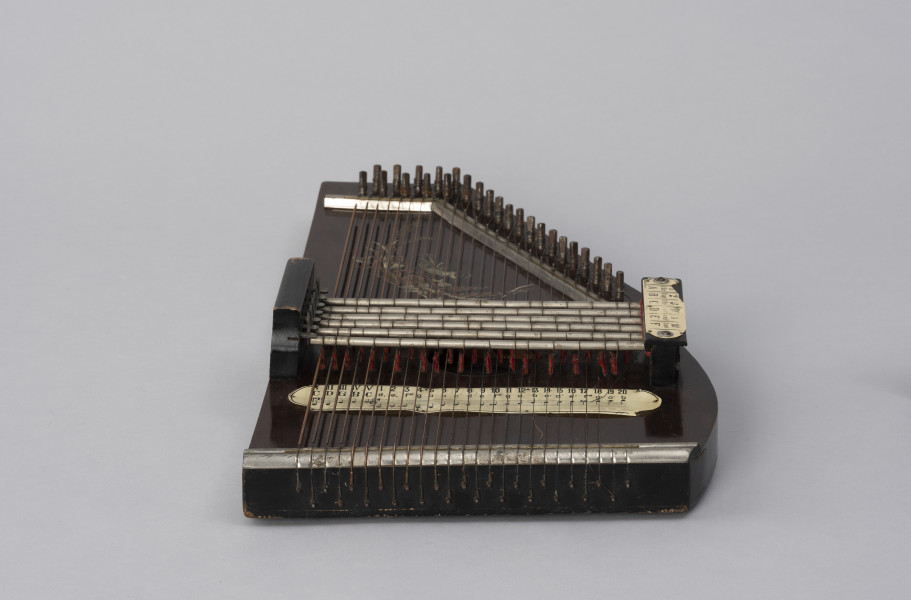 Cytra klawiszowa (autoharp) - ujęcie z dołu; Cytra klawiszowa (autoharp) z płaskim pudłem rezonansowym wykonanym z drewna polichromowanego o kształcie przypominającym trapez z jednym ściętym rogiem. Wzdłuż najkrótszego boku oraz boku odchodzącego od niego pod kontem rozwartym, szereg metalowych kołków, do których przymocowanych jest 25, diatonicznie strojonych strun, ułożonych od najgrubszej – z lewej – do najcieńszej z prawej strony instrumentu; ostatnie dwie są zerwane. Poniżej kołków znajdują się dwie, zestawione pod kątem rozwartym, wąskie, metalowe blaszki z niewielkimi bolcami, stanowiące wsparcie i gwarantujące właściwy rozstaw strun. Podobna blaszka zamontowana jest wzdłuż dolnej krawędzi instrumentu. Struny przechodzą przez znajdujące się w niej rowki, zaginają pod kątem 90 stopni i mocowane są w znajdujących się w bocznej ściance instrumentu bolcach, ułożonych naprzemiennie: wyżej – niżej. Bezpośrednio nad strunami, prostopadle względem nich, znajduje się sześć metalowych prętów zamontowanych pomiędzy dwoma drewnianymi mostkami. Osadzone na sprężynach pręty wprawiane są w ruch przez grającego za pomocą znajdujących się w ich górnej części przycisków. Powoduje to dociśnięcie do struny mocowanych na prętach klawiszy z filcowymi podkładkami. Na jednym z mostków, na przykręconej za pomocą dwóch śrub plakietce z papieru welinowego, znajdują się oznaczenia akordów. Druga plakietka, z zapisem nut w formie literowej i na pięciolinii oraz ciągiem cyfr rzymskich i arabskich, umieszczona jest bezpośrednio na pudle rezonansowym, poniżej mechanizmu klawiszowego. Cytra ozdobiona jest w górnej części kalkomanią przedstawiającą ptaszka, prawdopodobnie wróbla, siedzącego na ukwieconej gałązce.