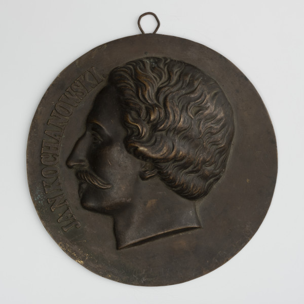 Medalion z wizerunkiem Jana Kochanowskiego - ujęcie z przodu; Okrągły medalion z dolutowanym uchem i wizerunkiem głowy męskiej w lewo. Głowa poety z krótkimi falującymi włosami i długimi wąsami. Z lewej strony napis JAN KOCHANOWSKI.