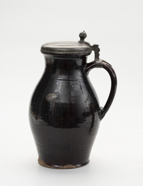 Dzban z pokrywą - ujęcie z przodu; Wysmukły dzban o lekko wyodrębnionej podstawie. Największa szerokość brzuśca przypada w połowie wysokości naczynia. Brzusiec łagodnym przewężeniem przechodzi w szeroką szyjkę; miejsce przejścia zaznaczone dookolną linią. Szyjka łagodnie przechodzi w wywinięty na zewnątrz wylew o zaoblonym rancie. Ucho szerokie, taśmowe, zaczepione pomiędzy dolną częścią szyjki, a  brzuścem poniżej najszerszej jego części. Na szczycie ucha obejma ze skierowaną pionowo do góry, ozdobnie profilowana kolumienką zaopatrzoną w zawias, służący do przymocowania pokrywy, szczelnie zamykającej wylew. Pokrywka z bolcem w postaci kolumienki zakończonej kopułką z małą kulką na szczycie, tworzące paluch do jej podnoszenia. Dzban  w całości pokryty szkliwem. Naczynie z wyraźnymi śladami naprawy: dziura w brzuścu zalepiona cynową, lutowana łatą, widoczne ślady po odrutowaniu, utrzymującym w całości pękniętą konstrukcję.