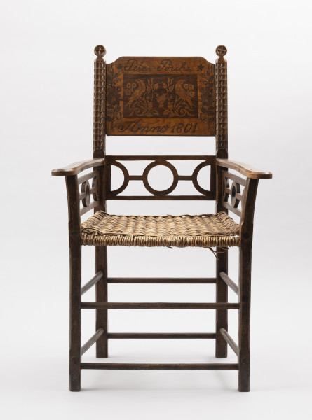 Fotel - ujęcie z przodu; Fotel - krzesło z podłokietnikami, wyrób rzemieślniczy stolarski, wykonawca nieznany. Mebel o konstrukcji ramowej z pionowo wznoszącym się oparciem. Nogi proste o przekroju prostokątnym, łączone dwiema toczonymi podpórkami, przednie z delikatnym wzorem geometrycznym wykonanym techniką snycerską. Siedzisko proste, wyplatane wikliną. Pomiędzy siedziskiem, dolną częścią oparcia i podłokietnikami ażurowo ozdobnie wycięta listwa ( motyw koła) będąca zarazem elementem konstrukcji. Podłokietniki na końcach lekko wywinięte na zewnątrz. Rama oparcia na obu listwach ze snycerską dekoracją w postaci stylizowanych kwiatów ( ?) i górnym zwieńczeniem w kształcie okręgu z rytym krzyżem. W ramę, w jej górnej części wpuszczona deska, jej górne krawędzie lekko wycięte na zewnętrznych rogach. Powierzchnia deski, z wyraźnie widoczną strukturą drewna, bogato zdobiona intarsją w dwóch odcieniach drewna. W środkowej części zaznaczony ciemniejszym kolorem drewna prostokąt, w środku którego symetrycznie umieszczone ptaki ( pawie ?)siedzące na stylizowanych gałęziach tulipanów i słoneczników (?). Nad prostokątem napis: Peter Buhr, pod nim: Anno 1801, po bokach motywy florystyczne.
