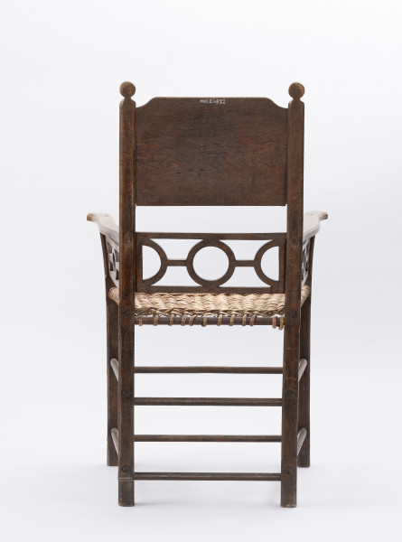 Fotel - ujęcie z tyłu; Fotel - krzesło z podłokietnikami, wyrób rzemieślniczy stolarski, wykonawca nieznany. Mebel o konstrukcji ramowej z pionowo wznoszącym się oparciem. Nogi proste o przekroju prostokątnym, łączone dwiema toczonymi podpórkami. Siedzisko proste, wyplatane wikliną. Pomiędzy siedziskiem, dolną częścią oparcia i podłokietnikami ażurowo ozdobnie wycięta listwa (motyw koła) będąca zarazem elementem konstrukcji. W ramę oparcia, w jej górnej części wpuszczona deska, jej górne krawędzie lekko wycięte na zewnętrznych rogach.