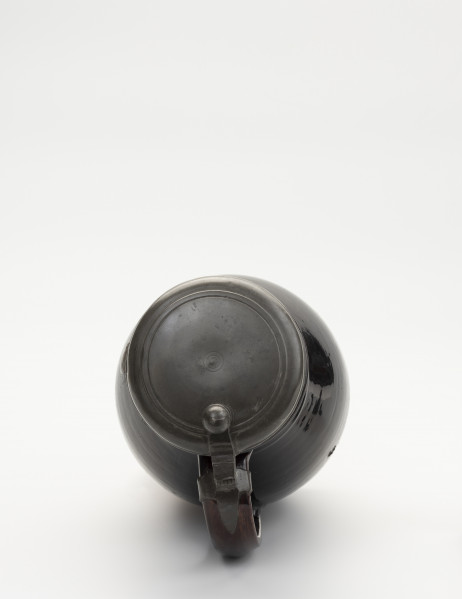 Dzban z pokrywą - ujęcie z góry; Ujęcie dzbana z góry. Największa szerokość brzuśca przypada w połowie wysokości naczynia. Ucho szerokie, taśmowe, zaczepione pomiędzy dolną częścią szyjki, a  brzuścem poniżej najszerszej jego części. Na szczycie ucha obejma ze skierowaną pionowo do góry, ozdobnie profilowana kolumienką zaopatrzoną w zawias, służący do przymocowania pokrywy, szczelnie zamykającej wylew. Pokrywka z bolcem w postaci kolumienki zakończonej kopułką z małą kulką na szczycie, tworzące paluch do jej podnoszenia. Na pokrywie proste liniowo-koncentryczne zdobienie uzyskane prawdopodobnie cyzelowaniem. Dzban  w całości pokryty szkliwem.