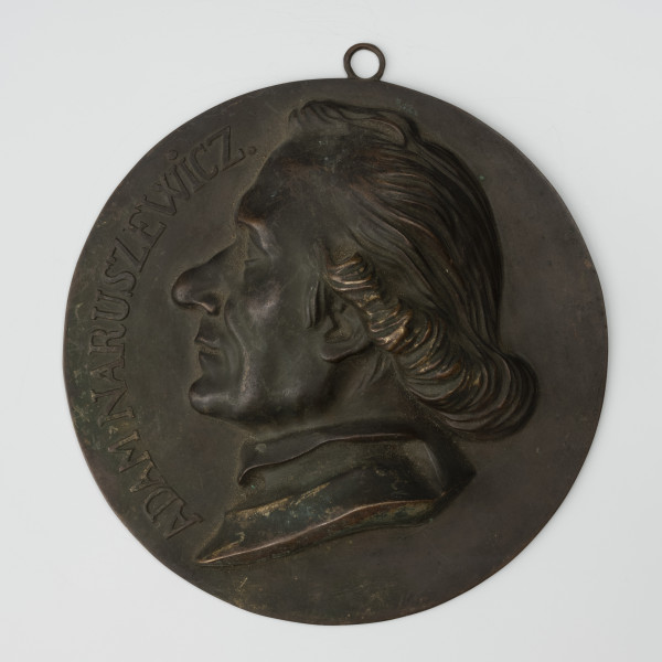 Medalion z wizerunkiem Adama Naruszewicza - ujęcie z przodu; Okrągły medalion z uchem i portretem męskim. Głowa mężczyzny zwrócona w prawo z włosami zaczesanymi do tyłu i podwiniętymi w luźny wałek. Z lewej strony napis: ADAM NARUSZEWICZ.