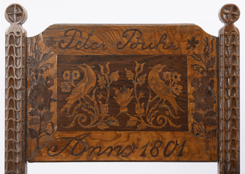 Fotel - detal; Rama oparcia na obu listwach ze snycerską dekoracją w postaci stylizowanych kwiatów i górnym zwieńczeniem w kształcie okręgu z rytym krzyżem. W ramę, w jej górnej części wpuszczona deska, jej górne krawędzie lekko wycięte na zewnętrznych rogach. Powierzchnia deski, z wyraźnie widoczną strukturą drewna, bogato zdobiona intarsją w dwóch odcieniach drewna. W środkowej części zaznaczony ciemniejszym kolorem drewna prostokąt, w środku którego symetrycznie umieszczone ptaki siedzące na stylizowanych gałęziach tulipanów i słoneczników (?). Nad prostokątem napis: Peter Buhr, pod nim: Anno 1801, po bokach motywy florystyczne.