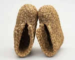 Buty słomiane - ujęcie z góry; Buty słomiane wykonane techniką plecionkarską. Z powróseł słomy wykonane buty o gładkiej podeszwie (bez obcasa), zabudowanej stopie. Cholewka z przodu rozchylona. Poszczególne elementy spirali warkocza  zszywane sznurkiem konopnym. Buty słomiane stosowane były do ocieplenia nóg w trakcie zimowych prac gospodarczo - rolniczych, jak np. w czasie młocki cepami.