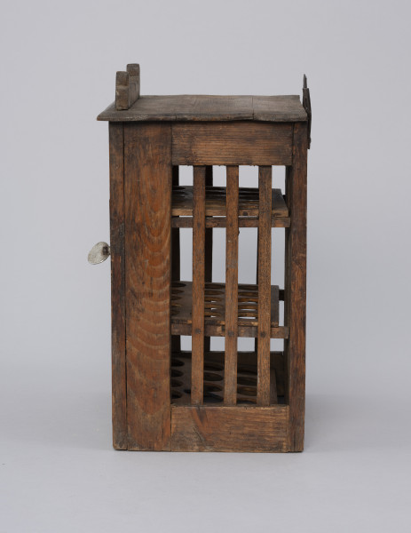 Szafka do przechowywania jajek - ujęcie z boku; Szafka do przechowywania jajek o konstrukcji ramowej w kształcie prostopadłościanu. Płycina ścianki bocznej wypełniona trzema pionowymi szczebelkami. Drzwiczki zamykane na zapadkę uruchamianą klamką. Wewnątrz trzy półki z wyciętymi otworami do wkładania jaj. Na desce zamykającej szafkę od góry przybita ozdobnie fazowana listwa.