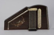 Cytra klawiszowa (autoharp) - ujęcie z góry; Cytra klawiszowa (autoharp) z płaskim pudłem rezonansowym wykonanym z drewna polichromowanego o kształcie przypominającym trapez z jednym ściętym rogiem. Wzdłuż najkrótszego boku oraz boku odchodzącego od niego pod kontem rozwartym, szereg metalowych kołków, do których przymocowanych jest 25, diatonicznie strojonych strun, ułożonych od najgrubszej – dolnej – do najcieńszej u góry instrumentu; ostatnie dwie są zerwane. Z prawej strony kołków znajdują się dwie, zestawione pod kątem rozwartym, wąskie, metalowe blaszki z niewielkimi bolcami, stanowiące wsparcie i gwarantujące właściwy rozstaw strun. Podobna blaszka zamontowana jest z prawej strony wzdłuż krawędzi instrumentu. Struny przechodzą przez znajdujące się w niej rowki, zaginają pod kątem 90 stopni i mocowane są w znajdujących się w bocznej ściance instrumentu bolcach, ułożonych naprzemiennie. Na poziomie otworu rezonansowego, bezpośrednio nad strunami, prostopadle względem nich, znajduje się sześć metalowych prętów zamontowanych pomiędzy dwoma drewnianymi mostkami. Osadzone na sprężynach pręty wprawiane są w ruch przez grającego za pomocą znajdujących się w ich górnej części przycisków. Na jednym z mostków, na przykręconej za pomocą dwóch śrub plakietce z papieru welinowego, znajdują się oznaczenia akordów. Druga plakietka, z zapisem nut w formie literowej i na pięciolinii oraz ciągiem cyfr rzymskich i arabskich, umieszczona jest bezpośrednio na pudle rezonansowym z prawej strony mechanizmu klawiszowego. Cytra ozdobiona jest z prawej strony kalkomanią przedstawiającą ptaszka, prawdopodobnie wróbla, siedzącego na ukwieconej gałązce.