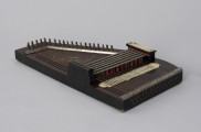 Cytra klawiszowa (autoharp) - ujęcie z drugiego skosu; Cytra klawiszowa (autoharp) z płaskim pudłem rezonansowym wykonanym z drewna polichromowanego o kształcie przypominającym trapez z jednym ściętym rogiem. Wzdłuż najkrótszego boku oraz boku odchodzącego od niego pod kontem rozwartym, szereg metalowych kołków, do których przymocowanych jest 25, diatonicznie strojonych strun, ułożonych od najgrubszej – z lewej – do najcieńszej z prawej strony instrumentu; ostatnie dwie są zerwane. Poniżej kołków znajdują się dwie, zestawione pod kątem rozwartym, wąskie, metalowe blaszki z niewielkimi bolcami, stanowiące wsparcie i gwarantujące właściwy rozstaw strun. Podobna blaszka zamontowana jest wzdłuż dolnej krawędzi instrumentu. Struny przechodzą przez znajdujące się w niej rowki, zaginają pod kątem 90 stopni i mocowane są w znajdujących się w bocznej ściance instrumentu bolcach, ułożonych naprzemiennie: wyżej – niżej. Bezpośrednio nad strunami, prostopadle względem nich, znajduje się sześć metalowych prętów zamontowanych pomiędzy dwoma drewnianymi mostkami. Osadzone na sprężynach pręty wprawiane są w ruch przez grającego za pomocą znajdujących się w ich górnej części przycisków. Powoduje to dociśnięcie do struny mocowanych na prętach klawiszy z filcowymi podkładkami. Na jednym z mostków, na przykręconej za pomocą dwóch śrub plakietce z papieru welinowego, znajdują się oznaczenia akordów. Druga plakietka, z zapisem nut w formie literowej i na pięciolinii oraz ciągiem cyfr rzymskich i arabskich, umieszczona jest bezpośrednio na pudle rezonansowym, poniżej mechanizmu klawiszowego. Cytra ozdobiona jest w górnej części kalkomanią.