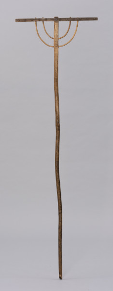 Grabie drewniane dwunastozębne - ujęcie z tyłu; Grabie drewniane o długim trzonku z okorowanego, lekko wygiętego kawałka drewna, ściętym na końcu, a bezpośrednio przed częścią pracującą, po jednej stronie opracowany płasko. U góry część pracująca składa się z prostokątnej listwy osadzonej prostopadle na trzonku. Konstrukcję grabi wzmacniają dwa rzędy łukowato wygiętych, okorowanych pałąków wykonanych prawdopodobnie z wierzbowej wici.