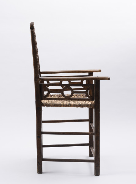 Fotel - ujęcie z boku; Fotel - krzesło z podłokietnikami. Mebel o konstrukcji ramowej z pionowo wznoszącym się oparciem. Nogi proste o przekroju prostokątnym, łączone dwiema toczonymi podpórkami, przednie z delikatnym wzorem geometrycznym wykonanym techniką snycerską. Siedzisko proste, wyplatane wikliną.