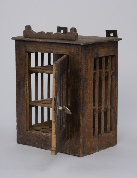 Szafka do przechowywania jajek - ujęcie ze skosu z prawej; Szafka do przechowywania jajek o konstrukcji ramowej w kształcie prostopadłościanu. Płycina jednoskrzydłowych drzwi oraz ścianki boczne  otwarte, wypełnione trzema pionowymi szczebelkami. Drzwiczki zamykane na zapadkę uruchamianą klamką. Wewnątrz trzy półki z wyciętymi otworami do wkładania jaj. Na desce zamykającej szafkę od góry przybita ozdobnie fazowana listwa. Plecy pełne, na górze przybite dwa haczyki do zawieszania.