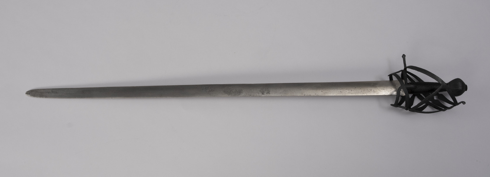 Pałasz typu schiavona - ujęcie z przodu; Pałasz z prostą, obusieczną głownią o przekroju soczewkowym. Na sztychu głowni przewiercony później otwór z pęknięciem. Trzon okrągły opleciony drutem. Jelec z koszem w typie szkieletowym składającym się z pionowego, wywiniętego na końcu pasa, od którego odchodzą dwie skośne boczne obręcze, biegnące do dolnej osłony pięści. Trzecia dolna obręcz przechodzi w środkową osłonę, nad którą jest jeszcze jedna równoległa. Do stabilizacji służą klamry, które odchodzą od dolnego wspornika bocznego i prowadzą do osłony poprzecznej. Z drugiej strony biegnie tylko jedna osłona pięści z jedną boczna obręczą, a u dołu zamocowany jest pierścień tzw. paluch. Głowica jest obła żłobkowana na powierzchni, z szerokim płaskim zakuciem od góry.