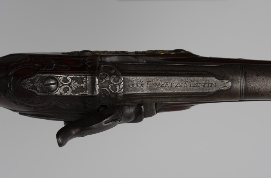 Para pistoletów kapiszonowych - detal; Fragment pistoletu kapiszonowego, pojedynkowego. Zamek kapiszonowy przerobiony ze skałkowych, zdobiony motywami roślinnymi i grawerowanym napisem: G.G. EWERTZ STETTIN.