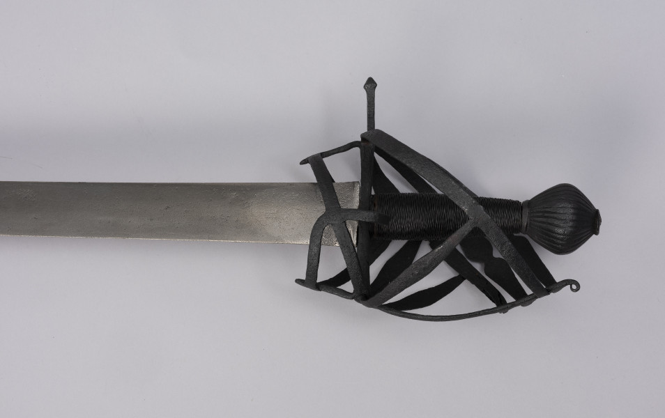 Pałasz typu schiavona - ujęcie detalu z przodu; Pałasz z prostą, obusieczną głownią o przekroju soczewkowym. Na sztychu głowni przewiercony później otwór z pęknięciem. Trzon okrągły opleciony drutem. Jelec z koszem w typie szkieletowym składającym się z pionowego, wywiniętego na końcu pasa, od którego odchodzą dwie skośne boczne obręcze, biegnące do dolnej osłony pięści. Trzecia dolna obręcz przechodzi w środkową osłonę, nad którą jest jeszcze jedna równoległa. Do stabilizacji służą klamry, które odchodzą od dolnego wspornika bocznego i prowadzą do osłony poprzecznej. Z drugiej strony biegnie tylko jedna osłona pięści z jedną boczna obręczą, a u dołu zamocowany jest pierścień tzw. paluch. Głowica jest obła żłobkowana na powierzchni, z szerokim płaskim zakuciem od góry.