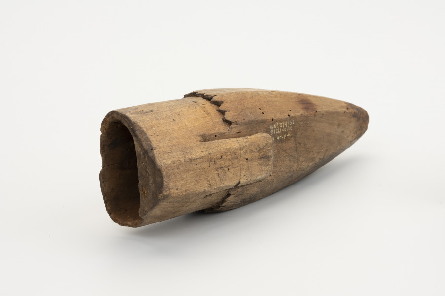 Pochewka na osełkę - ujęcie ze skosu; Pojemnik drewniany do przechowywania osełki z jednego kawałka ﻿drewna. Od górnej części o przekroju kwadratowym odprowadzony jest uchwyt służący do powieszenia przy pasku w trakcie prac polowych. Na wysokości uchwytu dookolnie wycięte ozdobne ząbki, poniżej których pochwa przybiera stożkowaty kształt. Środek wydrążony.