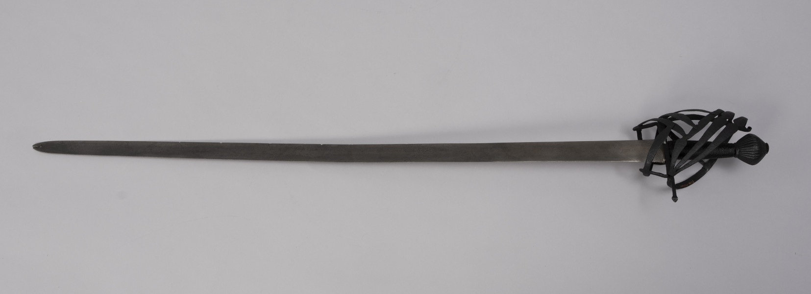 Pałasz typu schiavona - ujęcie z tyłu; Pałasz z prostą, obusieczną głownią o przekroju soczewkowym. Na sztychu głowni przewiercony później otwór z pęknięciem. Trzon okrągły opleciony drutem. Jelec z koszem w typie szkieletowym składającym się z pionowego, wywiniętego na końcu pasa, od którego odchodzą dwie skośne boczne obręcze, biegnące do dolnej osłony pięści. Trzecia dolna obręcz przechodzi w środkową osłonę, nad którą jest jeszcze jedna równoległa. Do stabilizacji służą klamry, które odchodzą od dolnego wspornika bocznego i prowadzą do osłony poprzecznej. Z drugiej strony biegnie tylko jedna osłona pięści z jedną boczna obręczą, a u dołu zamocowany jest pierścień tzw. paluch. Głowica jest obła żłobkowana na powierzchni, z szerokim płaskim zakuciem od góry.