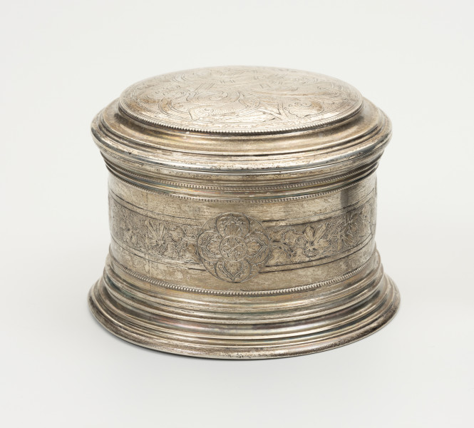 Puszka liturgiczna na komunikanty - ujęcie z tyłu; Okrągła puszka liturgiczna na komunikanty, wykonana z pozłacanego srebra. Wieczko okrągłe, z podwójnym, niezdobionym kołnierzem. Brzeg wieczka ze srebrnymi granulkami. Na ściance szeroki, okrągły pas ze zdobieniem kwietnym. Na środku pasa, w okręgu wychodzącym nieco poza ornamentowany pas, czteropłatkowy kwiat z okrągłym środkiem. Podstawa puszki okrągła, profilowana, złożona z koncentrycznych, stopniowanych obramowań.