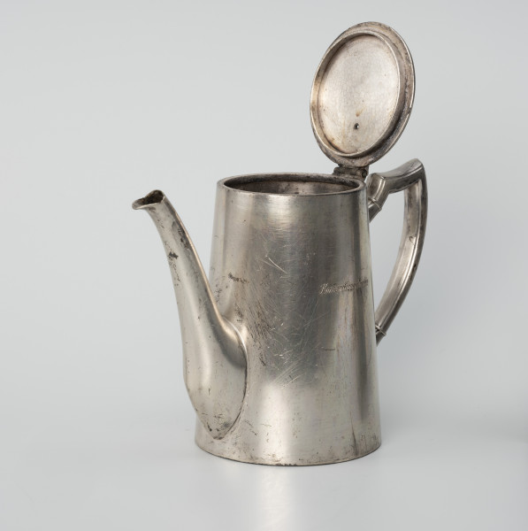 Dzbanuszek z zastawy Konzerthaus Stettin - ujęcie ze skosu z prawej; Dzbanuszek o prostej, zgeometryzowanej formie, bez wykształconej stopy. Korpus cylindryczny, zwężający się ku górze, zamykany płaską, okrągłą pokrywą. Dziubek osadzony w dolnej części korpusu, zwężający się ku górze, łagodnie wygięty, sięga wysokości pokrywy. Ucho o czworobocznym przekroju z profilem (rowkiem) na krawędziach, górą wyniesione do wysokości pokrywy, a następnie załamane, poprowadzone niemal poziomo i powtórnie załamane, a dalej delikatnie wygięte i łukiem wprowadzone do ścianki korpusu. Pokrywa - krążek z blachy na zawiasie powyżej ucha. Na korpusie grawerowany napis: Konzerthaus Stettin.