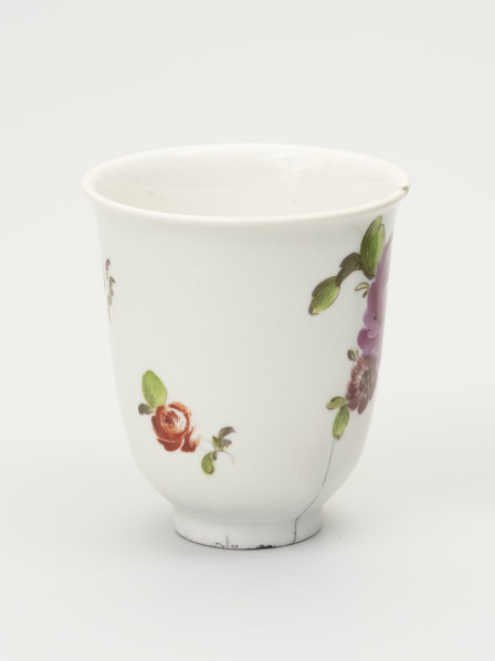 Czarka - ujęcie z boku; Smukła czarka na stopie o dzwonowatej formie z białej porcelany udekorowana malowanymi różnobarwnymi kwiatami. Przy kwiatach drobne zielone listki. U dołu z prawej pionowe pęknięcie.