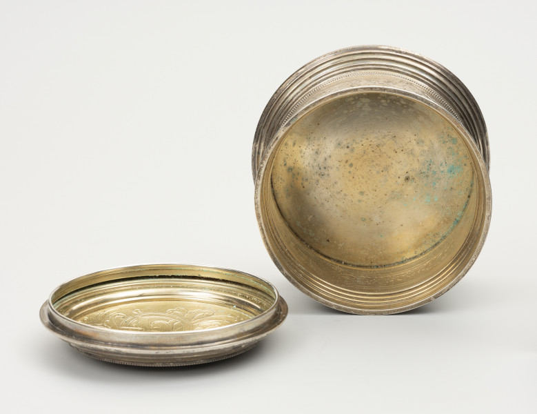 Puszka liturgiczna na komunikanty - ujęcie wewnątrz; Z prawej okrągła puszka liturgiczna na komunikanty, wykonana z pozłacanego srebra. Z lewej wieczko okrągłe, z podwójnym, niezdobionym kołnierzem. Brzeg wieczka ze srebrnymi granulkami. Podstawa puszki okrągła, profilowana, złożona z koncentrycznych, stopniowanych obramowań. Wnętrze gładkie z widocznymi korozjami.