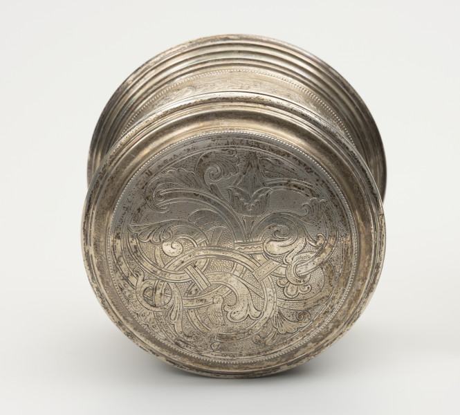 Puszka liturgiczna na komunikanty - ujęcie z góry; Okrągła puszka liturgiczna na komunikanty, wykonana z pozłacanego srebra. Wieczko okrągłe, z podwójnym, niezdobionym kołnierzem. Na wieczku ryty motyw roślinny: przenikające się pnącza roślinne, u dołu, po prawej stronie schematyczny ptak z profilu, ze złożonym skrzydłem i zaznaczonymi na nim piórami oraz okrągłym, pustym w środku okiem, małym dziobkiem, zwróconym w lewą stronę, a także schematycznymi kończynami. Brzeg wieczka ze srebrnymi granulkami. Podstawa puszki okrągła, profilowana, złożona z koncentrycznych, stopniowanych obramowań.