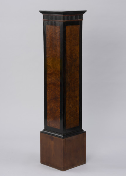 Postument dekoracyjny - ujęcie z czwartego skosu; Wolnostojący wysoki postument drewniany, fornirowany, z profilowaną krawędzią płyty wieńczącej i poszerzonym cokołem wykończonym profilowaną listwą. Pod płytą wieńczącą fryz z ciemnego, czarnego drewna z dekoracją w formie wąskich pionowych regularnych wyżłobień. Dzięki zastosowaniu różnych gatunków i barw forniru płyciny pionowych ścianek postumentu uzyskały ciemne, czarne obramienie w formie pasa przy krawędziach. Na płyciny ścianek bocznych wykorzystano jaśniejsze miodowe drewno o bogatym naturalnym wzorze czeczotowym.