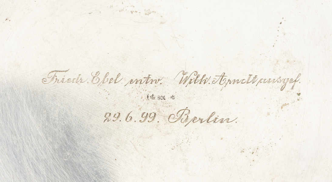 Puszka liturgiczna na komunikanty - detal; Fragment podstawy okrągłej puszki. Na środku napis ryty kursywą w dwóch liniach: Friedr. Ebel entn. Wilh. Arnolt (...)gef. | 29.6.99.Berlin. Pomiędzy linijkami wybite wklęsłe punce: ptak, zamknięta korona, cyfra 80C oraz ostatnia niezidentyfikowana punca.