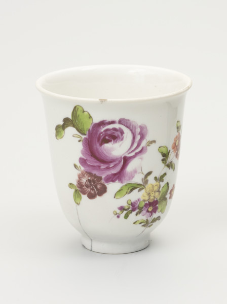 Czarka - ujęcie z przodu; Smukła czarka na stopie o dzwonowatej formie z białej porcelany udekorowana malowanymi różnobarwnymi piwoniami. Na pędzie głównym drobne kwiaty. Na lewym pędzie kwiaty fioletowe w tym jeden duży, pełny, rozwinięty. Z lewej strony od dołu widoczne pęknięcie idące w górę i zakończone tuż pod gałązką.