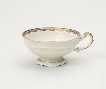 MNS/H/820 Filiżanka do herbaty - ujęcie z przodu; Duża filiżanka z białej porcelany dekorowana złoceniami. Czarka filiżanki okrągła w przekroju poziomym, szeroka na wysokości wylewu zwęża się w kierunku dna. Poniżej wylewu złocony ornament w formie ukośnego szrafowania ograniczonego od góry poziomą dookolną linią i od dołu ułożonymi na przemian C-kształtnymi elementami nawiązującymi do uproszczonego ornamentu małżowinowo-chrząstkowego. Na zewnętrznej ściance czarki oraz na stopce delikatne dookolne pasmo ornamentu plastycznego w formie spłaszczonych esownic i rozciągniętego motywu małżowinowo-chrząstkowego. Nawiązuje do niego również forma uszka ozdobionego pasmem złocenia na stronie grzbietowej, które przechodzi w cienką strużkę podkreślającą plastyczność ślimacznicy na profilu uszka, tuż przy łączeniu górnej jego części z czarką.