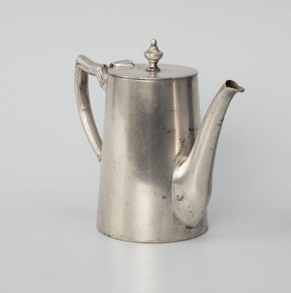 Dzbanuszek z zastawy Konzerthaus Stettin - ujęcie ze skosu z lewej; Dzbanuszek o prostej, zgeometryzowanej formie, bez wykształconej stopy. Korpus cylindryczny, zwężający się ku górze, zamykany płaską, okrągłą pokrywą z gałką w centrum. Dziubek osadzony w dolnej części korpusu, zwężający się ku górze, łagodnie wygięty, sięga wysokości pokrywy. Ucho o czworobocznym przekroju z profilem (rowkiem) na krawędziach, górą wyniesione do wysokości pokrywy, a następnie załamane, poprowadzone niemal poziomo i powtórnie załamane, a dalej delikatnie wygięte i łukiem wprowadzone do ścianki korpusu. Pokrywa - krążek z blachy na zawiasie powyżej ucha; uchwyt umocowany centralnie, w formie kielichowej gałki zwieńczonej kulką.