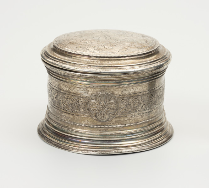 Puszka liturgiczna na komunikanty - ujęcie z boku; Okrągła puszka liturgiczna na komunikanty, wykonana z pozłacanego srebra. Wieczko okrągłe, z podwójnym, niezdobionym kołnierzem. Na wieczku ryty motyw roślinny. Brzeg wieczka ze srebrnymi granulkami. Na ściance szeroki, okrągły pas ze zdobieniem kwietnym. Na środku pasa, w okręgu wychodzącym nieco poza ornamentowany pas, czteropłatkowy kwiat z okrągłym środkiem. Podstawa puszki okrągła, profilowana, złożona z koncentrycznych, stopniowanych obramowań.