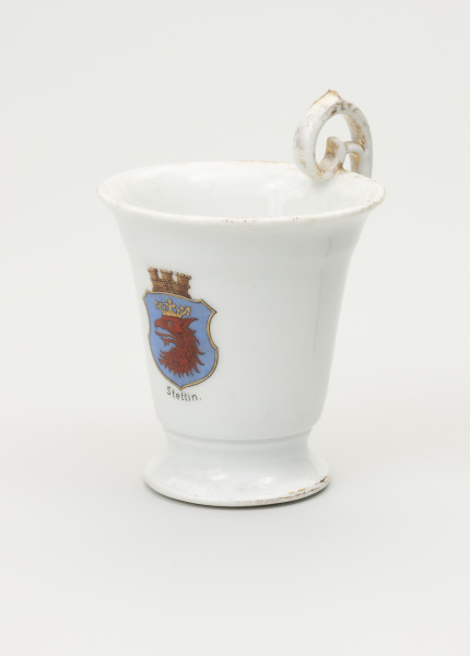 Filiżanka - ujęcie z boku; Filiżanka z grubej, białej porcelany w kształcie odwróconego dzwonu. Czara wysoka, łagodnie rozszerzająca się ku górze, z wylewem wywiniętym na zewnątrz. Stopa niska, okrągła, rozpłaszczona na zewnątrz, z pierścieniem na styku z czarą. Uszko wysokie, wyniesione ponad poziom wylewu w kształcie zwiniętego zwieńczenia pastorału; doklejone do wnętrza jej wylewu. Czara ozdobiona w centrum, po przeciwnej stronie uszka, malowaną naszkliwnie barwną tarczą z herbem Szczecina - profil głowy gryfa w koronie. Tarczę z bocznymi wcięciami wieńczy corona muralis a pod herbem napis Stettin. Ślady złoceń wokół wylewu od środka czary, na brzegu stopy oraz na uchwycie.